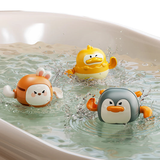 Three colorful wind-up swimming bath toys shaped like a bunny, duck, and penguin are splashing in a bathtub filled with water. The toys have cartoonish expressions and are actively moving, creating ripples and water splashes around them.