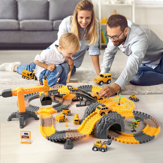 Happy family playing together with a large construction-themed toy car track set in the living room. The playset includes winding roads, tunnels, bridges, and a working crane, along with multiple yellow construction vehicles such as trucks, jeeps, and excavators. Road signs and palm tree decorations complete the realistic construction scene, encouraging imaginative play and parent-child interaction.