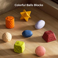 A collection of eight colorful sensory balls and blocks arranged on a wooden surface. The toys come in various shapes—cylinder, cube, sphere, star, and oval—and feature diverse textures such as bumps, ridges, and patterns for tactile stimulation. The text 