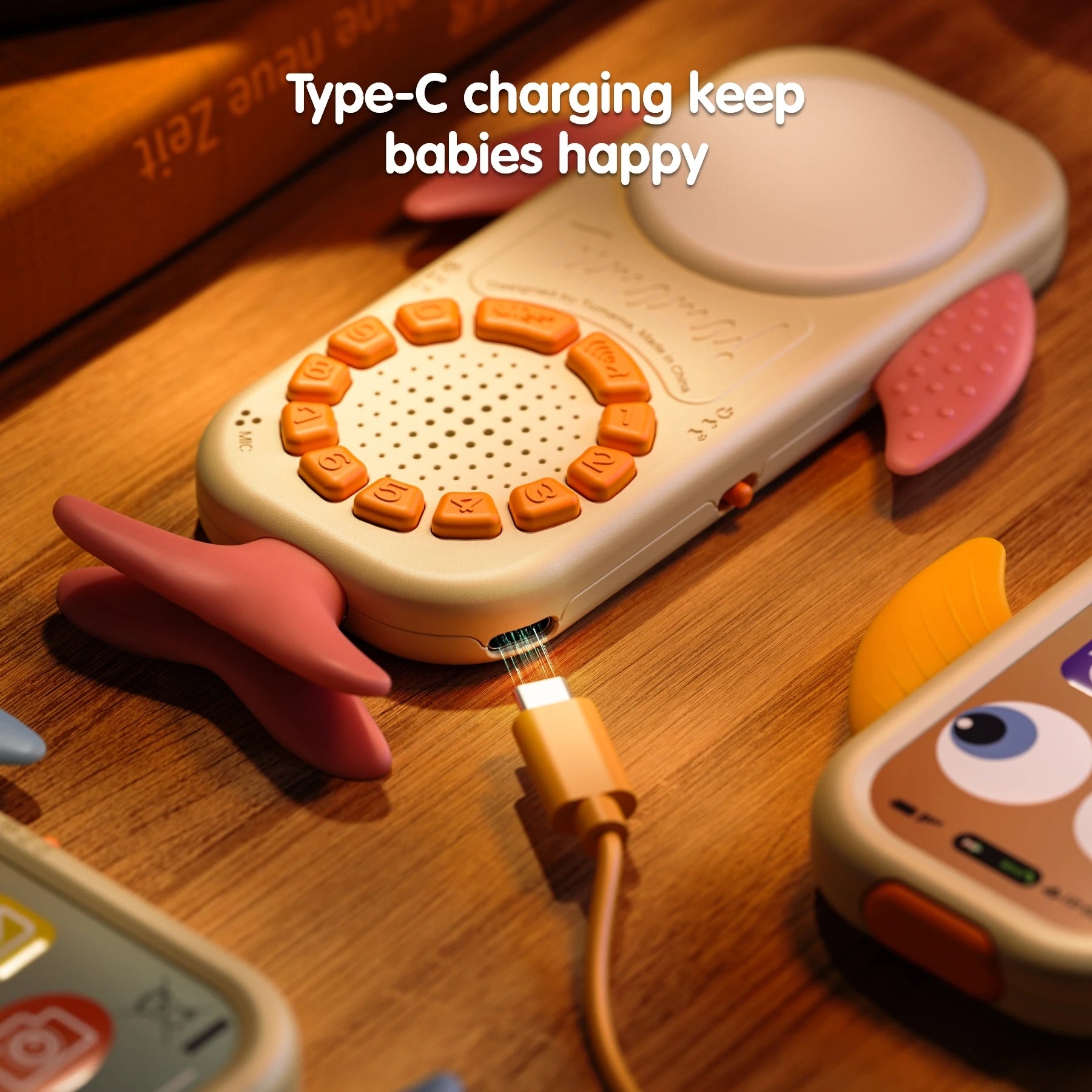 Close-up of a whale-shaped baby toy phone with pink fins being charged with a Type-C cable on a wooden surface. Text at the top reads “Type-C charging keep babies happy.”
