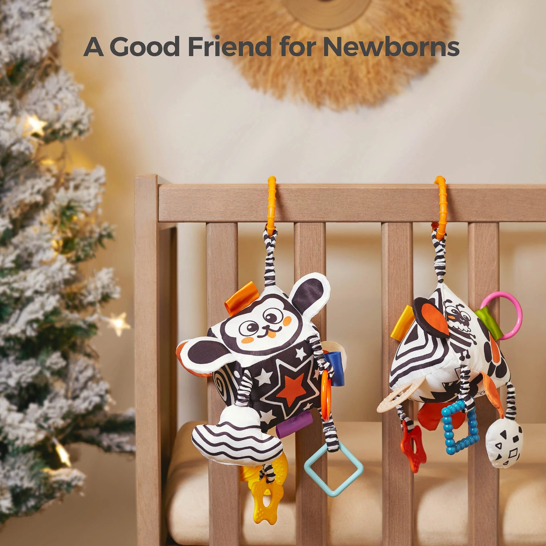 Two black and white high-contrast sensory cube toys hanging from a wooden crib rail. Each toy features colorful tags, teethers, rattles, and plush animal faces, designed to stimulate newborn vision and keep babies engaged. A Christmas tree with lights is visible in the background, creating a cozy nursery atmosphere.