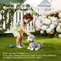 Child interacting with lifelike plush puppy toy outdoors, giving voice command “Sit down” as the toy sits on grass and wags its tail beside blooming hydrangeas and a white picket fence