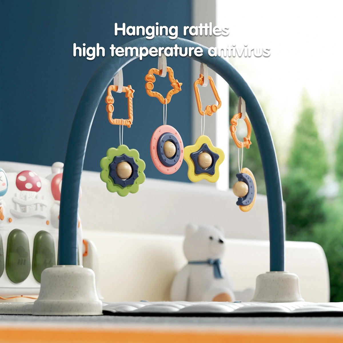 Close-up of a baby gym with a blue arch holding multiple colorful hanging rattles in star and circular shapes. A white bear plush toy is in the background, and a musical keyboard panel with cartoon faces is visible on the left.