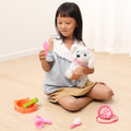 Smiling young girl sitting on the floor holding a white plush dog toy dressed in pink, while examining a toy mirror surrounded by pretend pet grooming accessories including a food bowl, bone, brush, hairdryer, and stethoscope.