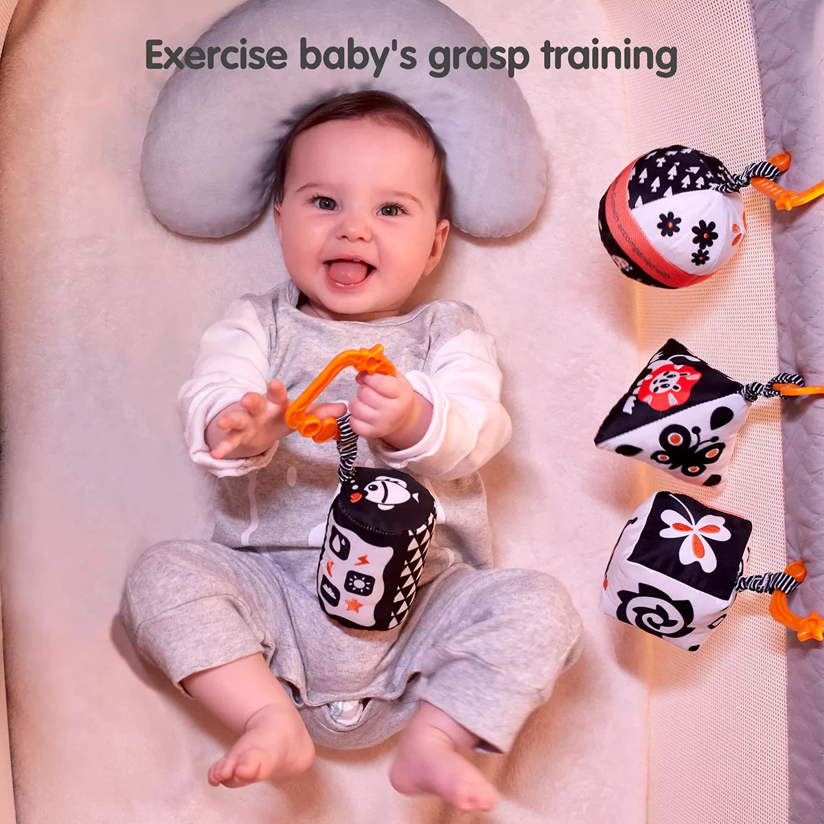 A smiling baby lying on a soft cushion, holding a black and white high-contrast rattle toy with an orange clip. Several similar toys are placed beside the baby. Designed to enhance visual development and grasping skills, these toys support early sensory and motor training.